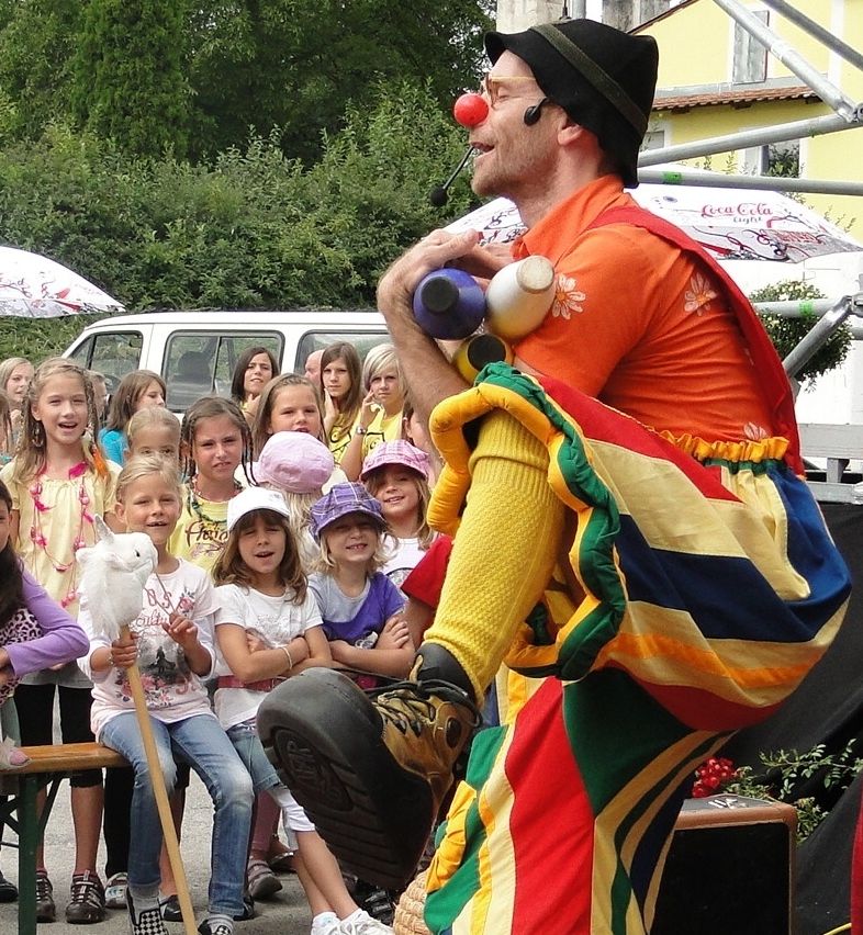Ein Clown mit roter Nase unterhält eine Gruppe von Kindern, die ihm aufmerksam zuschauen. Der Clown hält eine Flasche und einen Ballon.