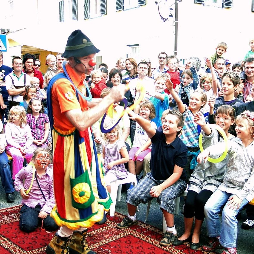 Ein Clown tritt vor einer Menge von Kindern und Erwachsenen auf, einige Kinder halten gelbe Ringe.