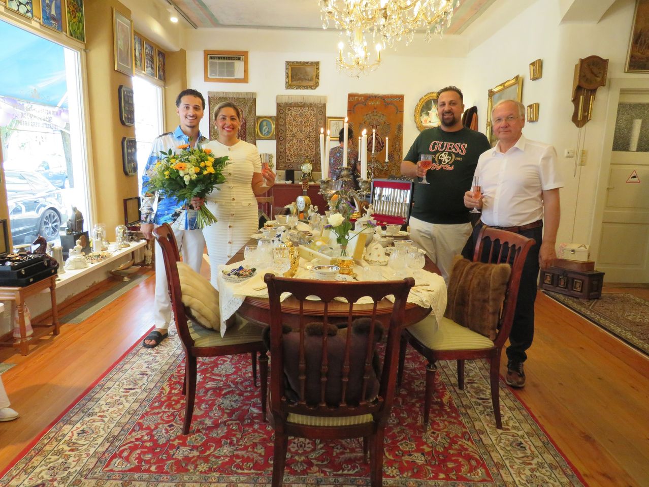 Four people are standing around a dining table in a well-decorated room, with a woman holding flowers and a man holding a wine glass. The table is set with a white tablecloth, flowers, and candles.
