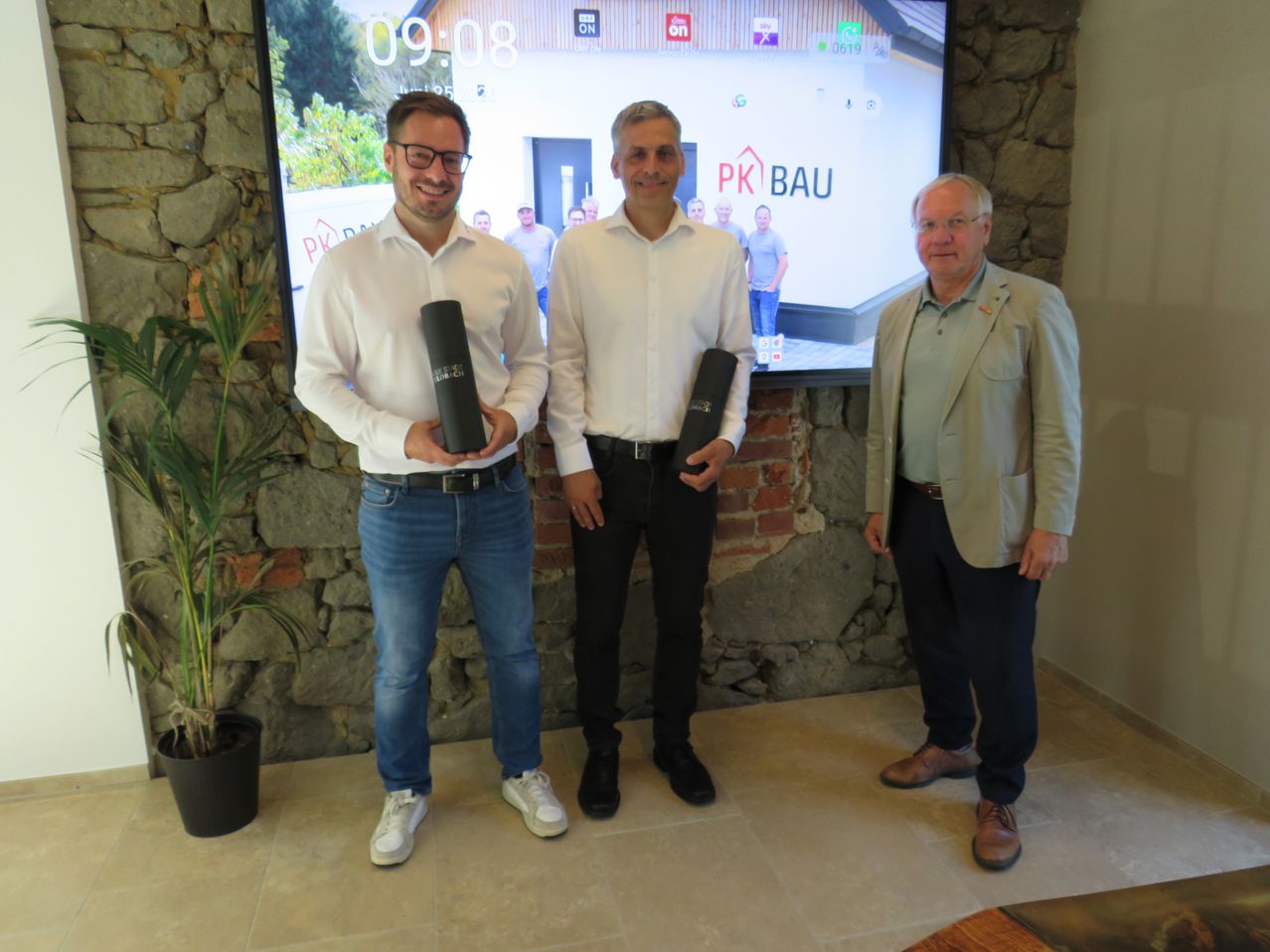 Three men stand in front of a stone wall, holding black cylindrical objects. The man on the left wears glasses and a belt. The man in the middle wears a watch and holds a black object. The man on the right wears a suit and glasses. Behind them is a large screen displaying a house.