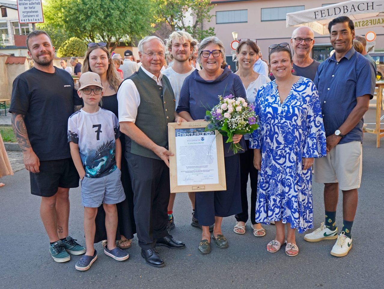 Eine Gruppe von Menschen, darunter eine Frau mit einem Blumenstrauß und einem gerahmten Zertifikat, steht vor einem Gebäude.