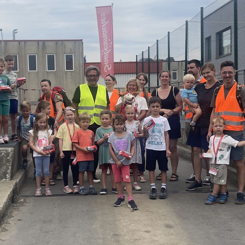 A group of children and adults wearing orange vests are standing outside a building, holding boxes and smiling for a photo.