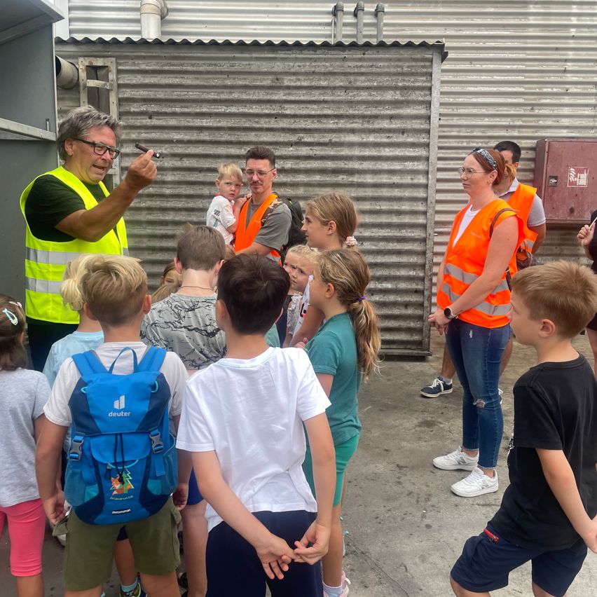 A group of children and adults, some wearing safety vests, are gathered around a man who is holding a pen and speaking to them. The setting appears to be an outdoor area with a corrugated metal wall in the background.