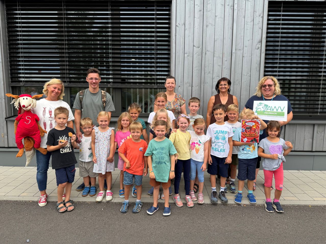 A group of children and adults pose for a photo in front of a building, some holding books and one holding a sign.