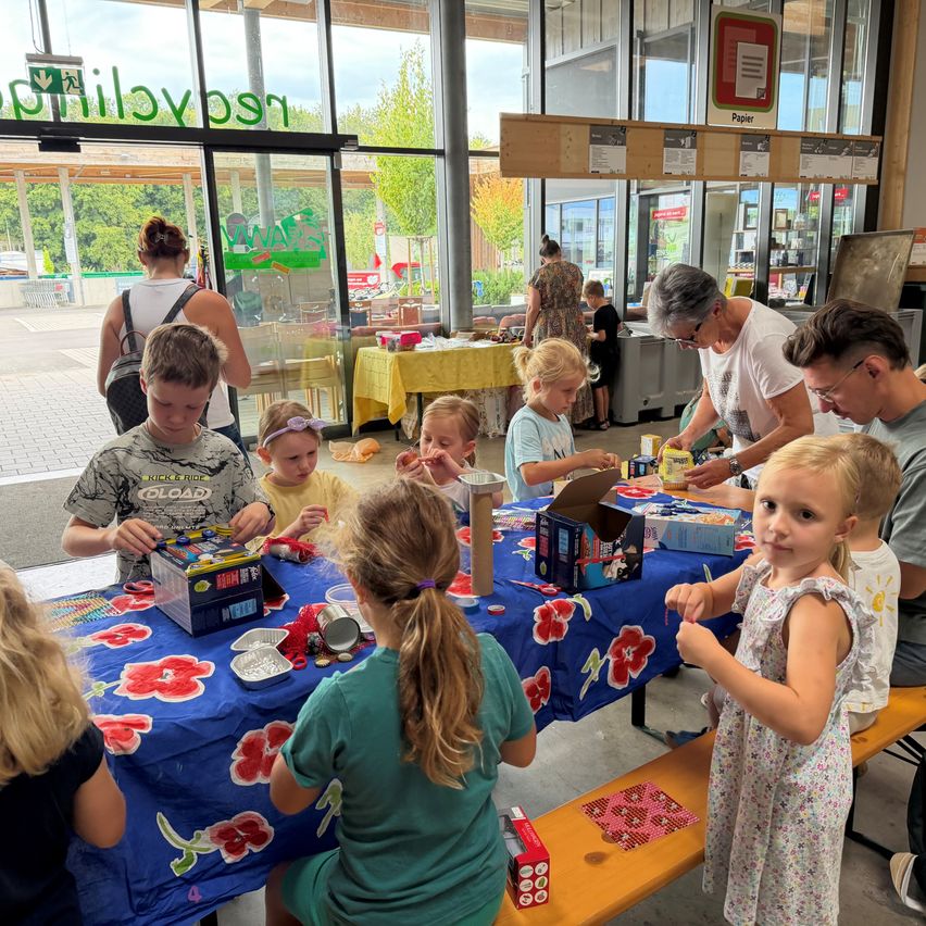 A group of children and adults are gathered around a table, engaged in a craft activity. The table is covered with a blue floral tablecloth, and various craft materials are spread out. A woman in glasses is assisting the children. In the background, there are glass doors and a sign.