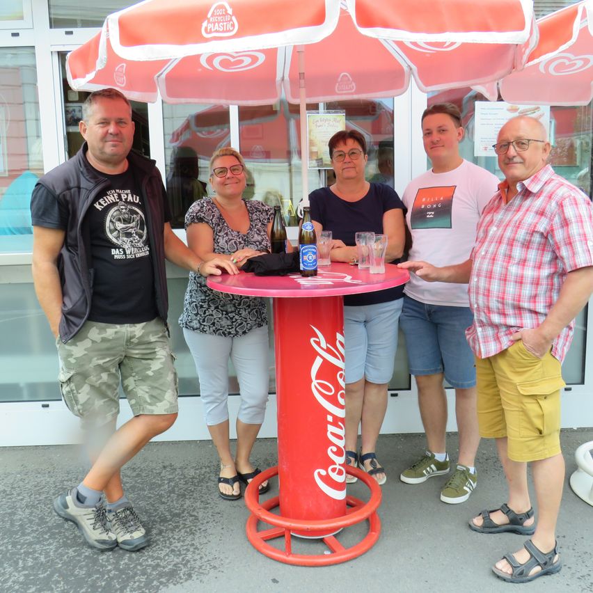 Five people stand around a Coca-Cola branded table, smiling for a photo. The table has a Coca-Cola bottle as its centerpiece, and they are under a red umbrella.