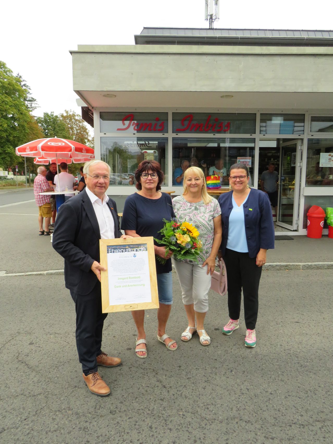 Four people stand in front of a store named 'Imbiss' with one man holding a certificate. Behind them, people are sitting at tables under an umbrella.