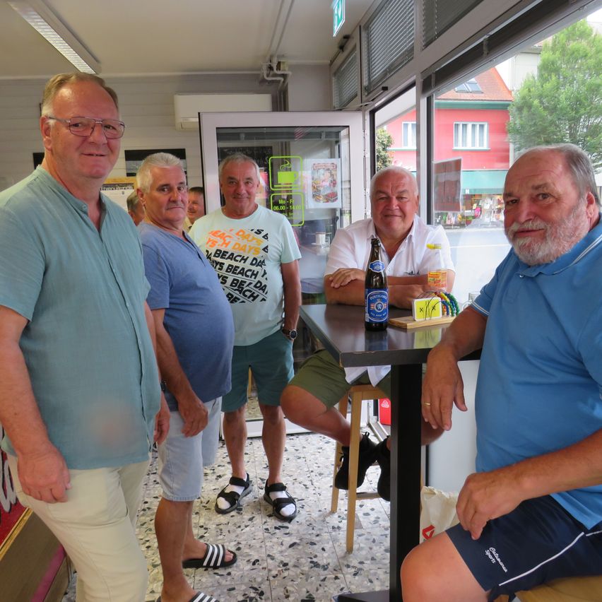Five men are standing and sitting in a restaurant, smiling and posing for a photo. One man is sitting on a stool with a beer bottle on the table.