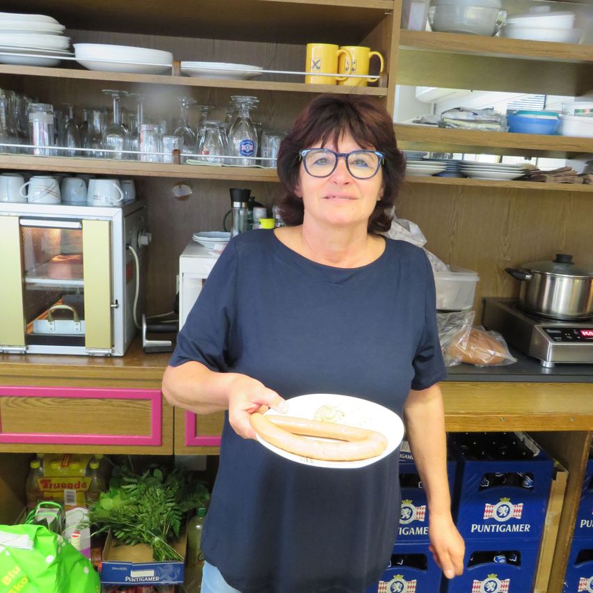 A woman in a blue shirt stands in a kitchen, holding a plate with two sausages. Behind her, shelves are stocked with plates, glasses, and kitchenware. A microwave and a stove are visible in the background.