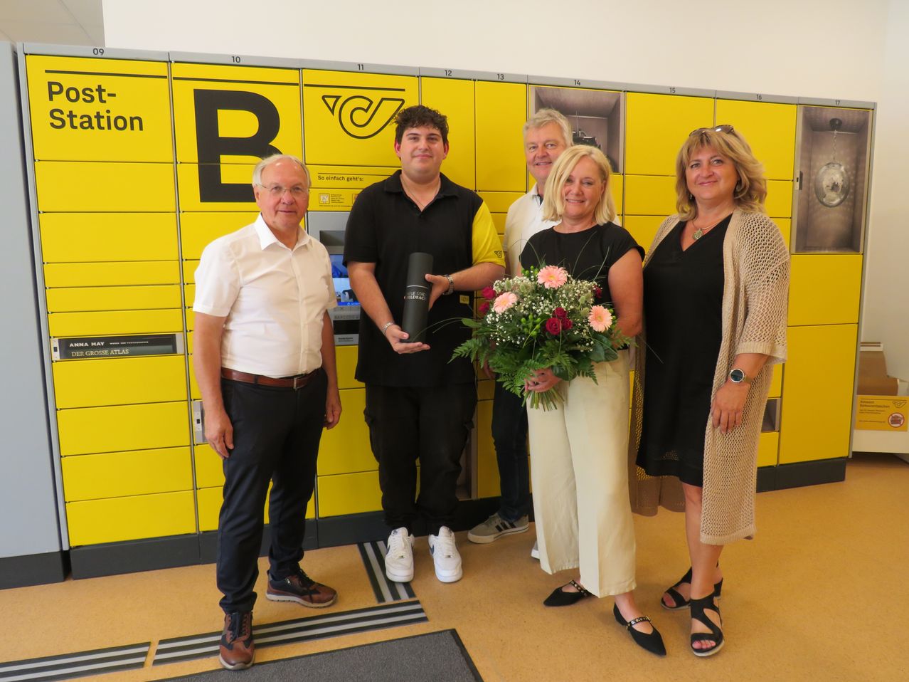 Fünf Personen stehen vor einer gelben Schließfachwand und lächeln für ein Foto. Der Mann links hält einen Blumenstrauß.