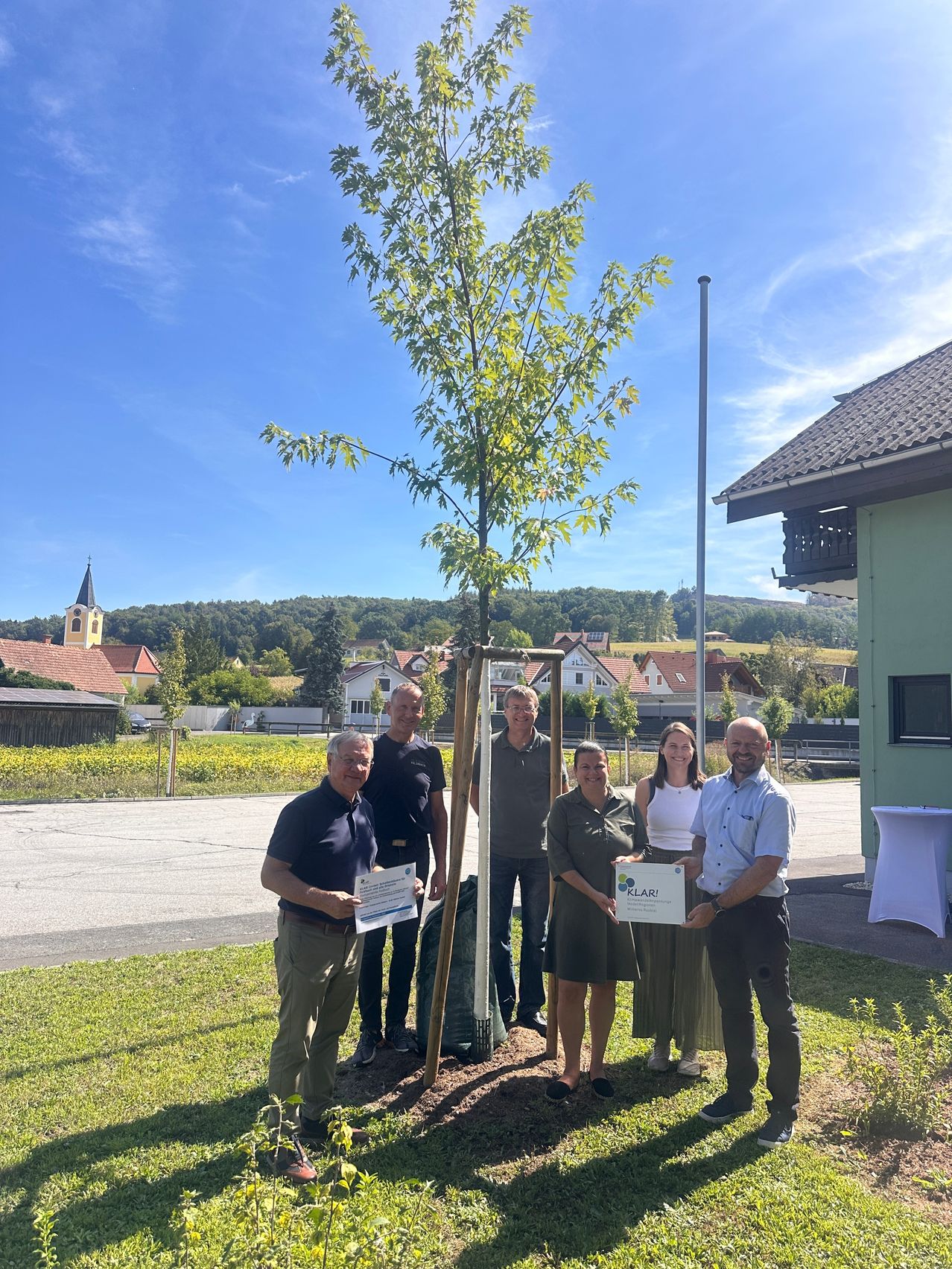 A group of people stand around a newly planted tree, holding certificates and smiling for the camera. The setting is outdoors with a house and village in the background.