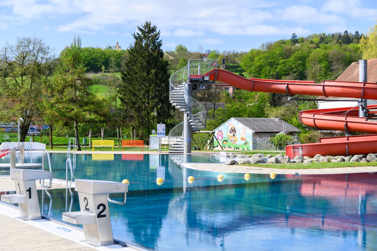 Ein Freibad mit einer Wendeltreppe, die zu einer roten Wasserrutsche führt. Der Pool ist von Grün umgeben und hat eine gelbe Bank auf der linken Seite.