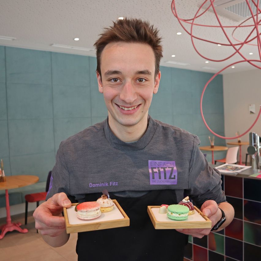 Ein lächelnder Mann in einem grauen T-Shirt hält zwei kleine Tabletts mit bunten Macarons in einem modernen Café.