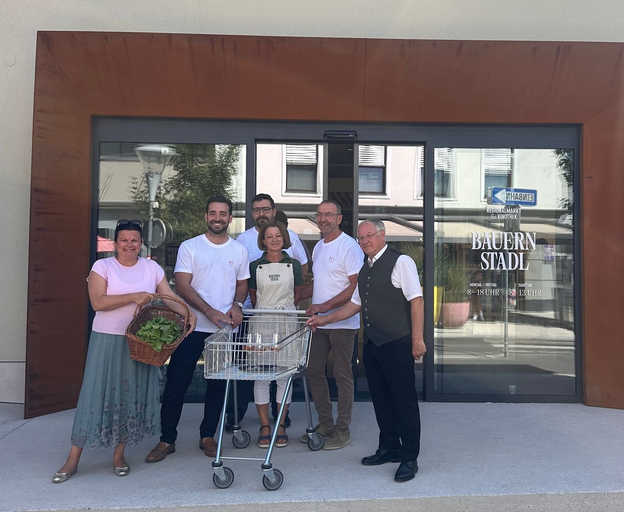 Eine Gruppe von Menschen steht vor einem Geschäft, lächelt und posiert für ein Foto. Sie halten einen Korb und einen Einkaufswagen. Das Geschäft hat Glastüren und ein Schild, das 'Bauern Stadt' steht.