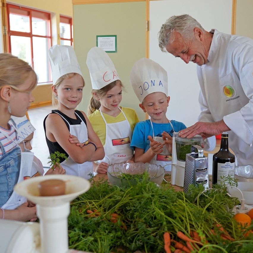 A chef is instructing a group of kids in a kitchen, who are wearing chef hats and aprons. They are preparing food, with carrots, parsley, and other vegetables on the table.