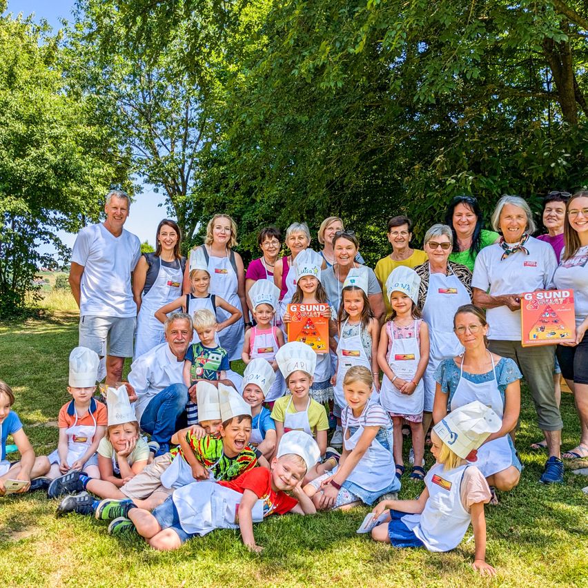 A group of adults and children are posing for a photo in a grassy area under trees. They are wearing aprons and chef hats. Some of them are holding books.