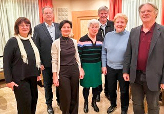 A group of people stands together in a room with beige walls, smiling for a photograph. They wear formal attire, including suits, scarves, and dresses. Behind them, a white curtain and a red curtain are visible.