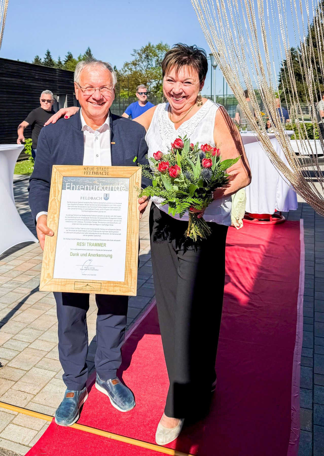 Ein Mann und eine Frau stehen zusammen und halten ein Zertifikat und einen Blumenstrauß auf einem roten Teppich. Der Mann trägt eine Brille und einen Anzug, die Frau einen Halsschmuck und Ohrringe. Dahinter sind Tische und ein Gehweg.