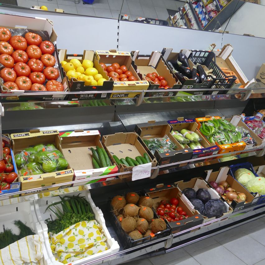 Ein Supermarktregal mit verschiedenen Gemüsen und Früchten in Kisten. Tomaten, Gurken, Auberginen und Zitronen gehören zu den ausgestellten Artikeln. Einige Kisten haben Preisschilder.