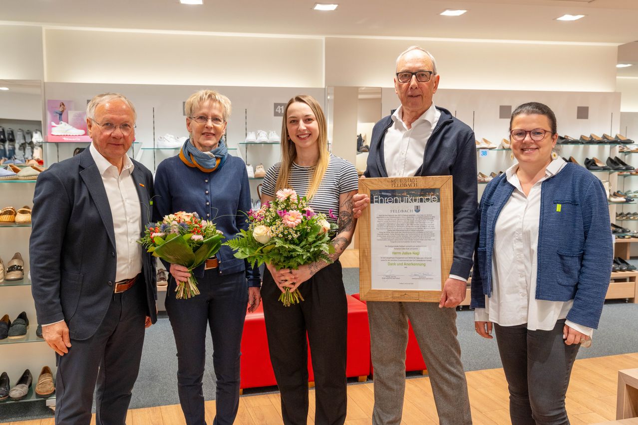 Fünf Personen posieren mit Blumen und einem gerahmten Zertifikat in einem Geschäft. Eine Frau in der Mitte lächelt mit einem Tattoo auf ihrem Arm. Ein Mann rechts trägt eine Brille und eine Jacke.