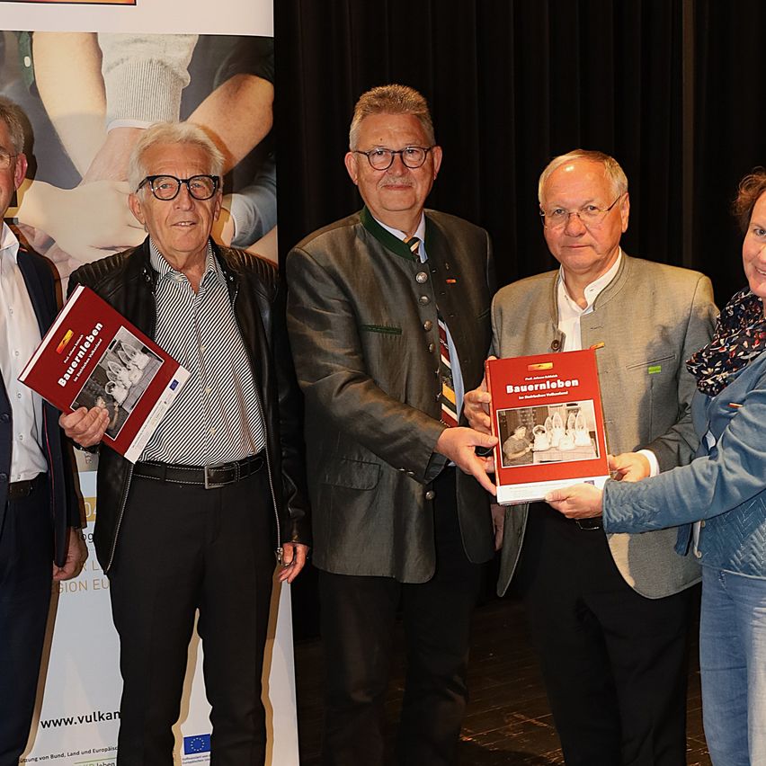 Vier Männer stehen in einem Raum und halten ein rotes Buch, möglicherweise eine Anerkennungszeremonie. Der Mann auf der linken Seite trägt eine schwarze Jacke. Der zentrale Mann trägt eine graue Jacke und eine Krawatte. Der Mann auf der rechten Seite trägt einen grauen Mantel.