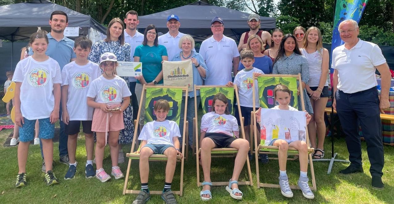 A group of adults and children are posing for a photo on a sunny day, with some holding a check and a bag. They are sitting on deck chairs on a grassy field.