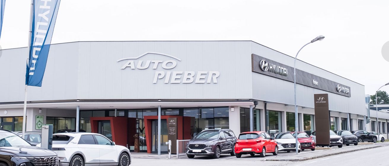 Ein weißes Gebäude mit AUTO PIEBER Schild über einem Hyundai-Händler. Mehrere Autos sind vor dem Gebäude geparkt.