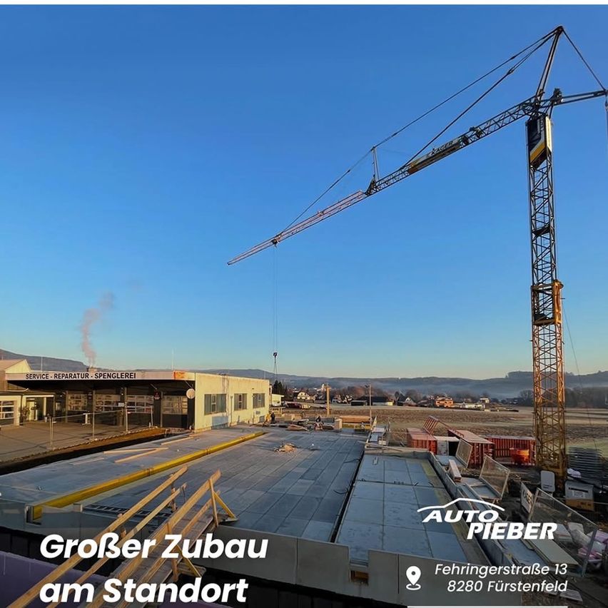A construction site with a large crane lifting materials. The image shows a building with the words 'Service-Reparatur-Spenglerei' and another building under construction. It includes a crane, a building under construction, and a clear blue sky.