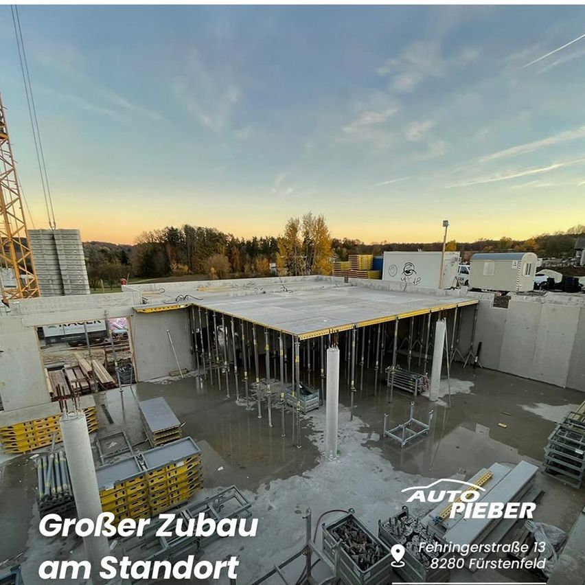 Construction site with a large building under construction. A crane is present. The site is surrounded by trees. The ground is wet.