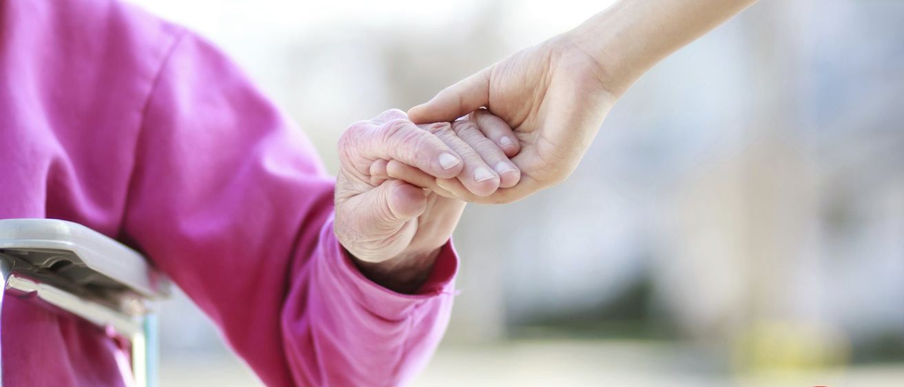 Eine ältere Person wird von einer anderen Person gehalten, beide tragen rosa lange Ärmel, möglicherweise in einer Pflegeeinrichtung. Ein grünes Logo ist in der unteren rechten Ecke sichtbar.