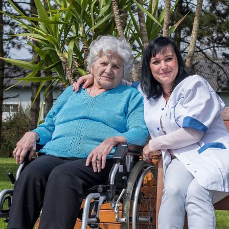 Eine ältere Frau im Rollstuhl sitzt neben einer lächelnden Frau in einer weißen Uniform, möglicherweise eine Krankenschwester, in einem Garten.