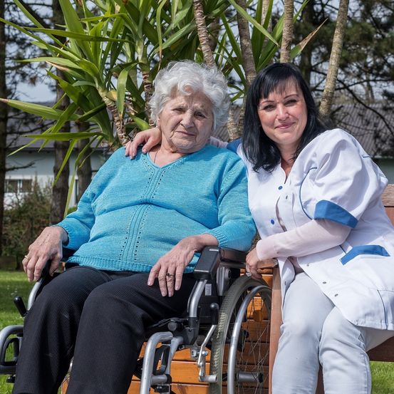Eine ältere Frau im Rollstuhl sitzt neben einer lächelnden Frau in einer weißen Uniform, möglicherweise eine Krankenschwester, in einem Garten.