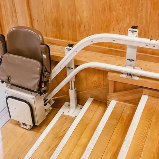 Ein Treppenlift mit braunem Sitz ist auf einer hölzernen Treppe positioniert, ausgestattet mit weißen Handläufen und einer hölzernen Bank an der Seite.