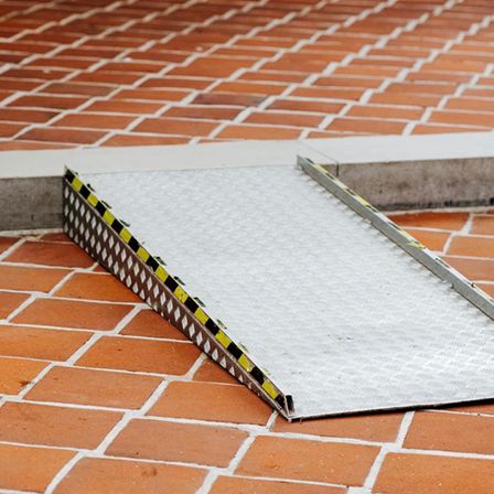 Eine graue Metallrampe mit schwarzen und gelben Streifen liegt auf einem roten Ziegelboden. Die Rampe ist in einem Winkel positioniert, möglicherweise für Barrierefreiheit.