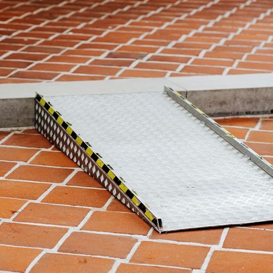 Eine graue Metallrampe mit schwarzen und gelben Streifen liegt auf einem roten Ziegelboden. Die Rampe ist in einem Winkel positioniert, möglicherweise für Barrierefreiheit.
