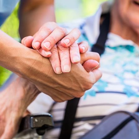 Eine Person im Rollstuhl hält die Hand einer anderen Person, beide zeigen Alterserscheinungen. Die Person im Rollstuhl trägt ein gestreiftes Hemd.