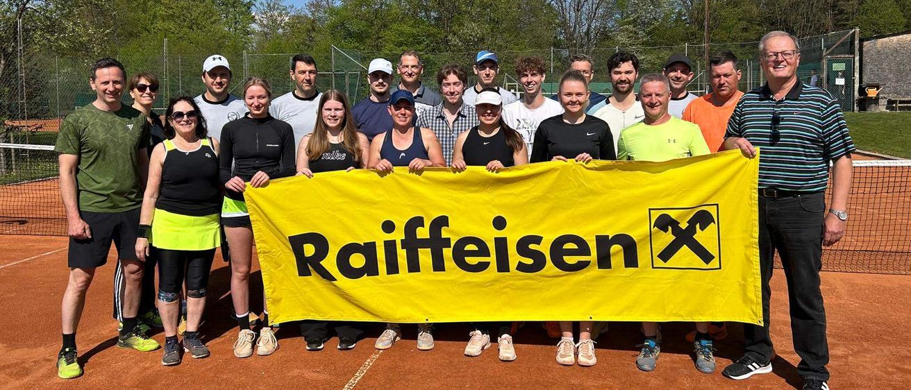 Eine Gruppe von Tennisspielern steht auf einem Platz und hält ein gelbes Banner mit dem Wort Raiffeisen. Sie lächeln und posieren für ein Foto. Dahinter ist ein Zaun und einige Bäume.