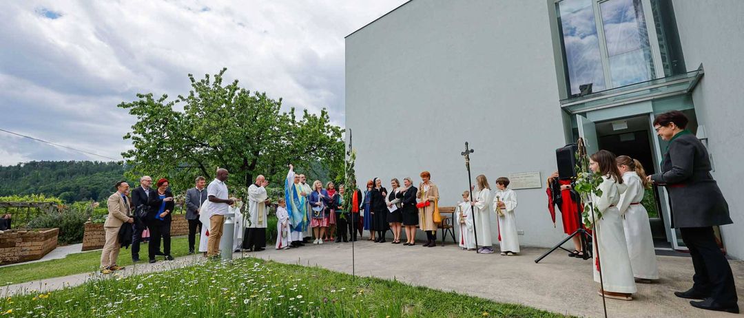 Eine Gruppe von Menschen in weißen Gewändern steht vor einem Kirchengebäude. Einige halten Blumen, während andere einem Lautsprecher zuhören. Das Gebiet ist von Gras und Bäumen umgeben.