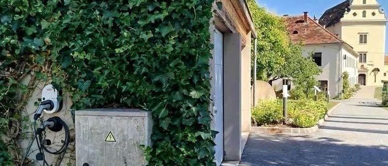 Eine Ladestation für Elektrofahrzeuge befindet sich in der Nähe eines mit Efeu bedeckten Gebäudes. Dahinter führt eine Einfahrt zu einem historischen Gebäude mit Turm.