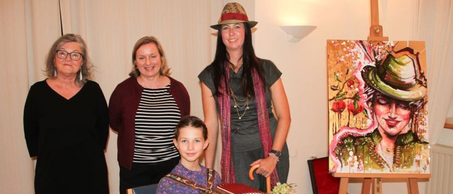 Vier Frauen und ein junges Mädchen posieren für ein Foto in einem Raum. Das Mädchen hält ein Akkordeon. Hinter ihnen steht ein Staffelei mit einem Gemälde einer Frau in einem Hut.