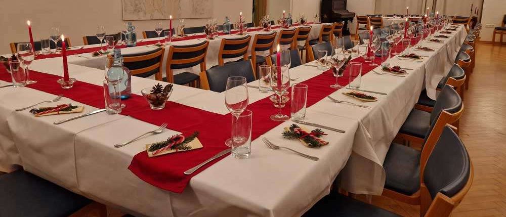 Ein langer Esstisch, der für einen besonderen Anlass gedeckt ist, mit roten Tischläufern, Weingläsern und Kerzen. Der Raum hat gewölbte Decken und ein großes Gemälde.