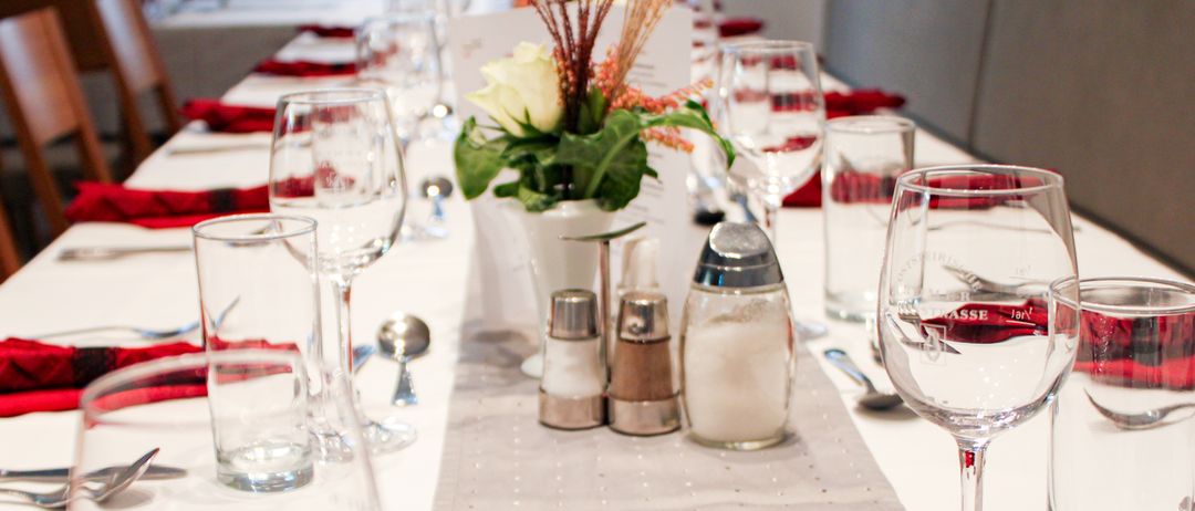 Ein langer Esstisch ist für eine formelle Veranstaltung gedeckt. Es gibt mehrere Weingläser, eine Blumenmitte und Gewürzshaker. Rote Servietten sind auf dem Tisch gefaltet.