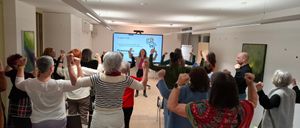 Eine Gruppe von Frauen ist in einem Raum versammelt und nimmt an einem Workshop teil. Eine Frau in der Mitte hält eine Präsentation auf einem Bildschirm, und andere heben ihre Hände.