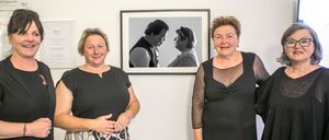 Drei Frauen stehen vor einem gerahmten Porträt. Das Porträt zeigt einen Mann und eine Frau in Schwarz-Weiß. Die Frauen lächeln und scheinen sich in einer Kunstgalerie zu befinden.