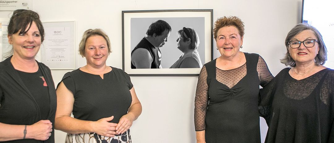 Drei Frauen stehen vor einem gerahmten Porträt. Das Porträt zeigt einen Mann und eine Frau in Schwarz-Weiß. Die Frauen lächeln und scheinen sich in einer Kunstgalerie zu befinden.