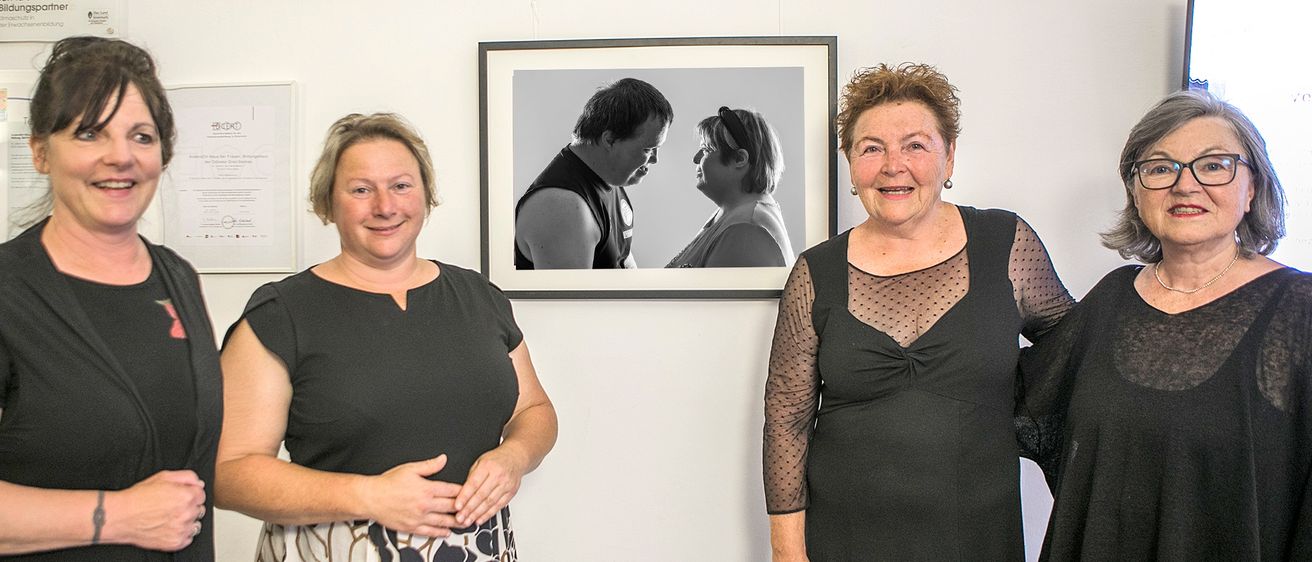 Drei Frauen stehen vor einem gerahmten Porträt. Das Porträt zeigt einen Mann und eine Frau in Schwarz-Weiß. Die Frauen lächeln und scheinen sich in einer Kunstgalerie zu befinden.