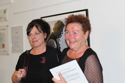Zwei erwachsene Frauen lächeln und posieren für ein Foto in einer Kunstgalerie. Eine Frau hält ein Dokument und einen Stift. Hinter ihnen befindet sich ein gerahmtes Foto einer Person.