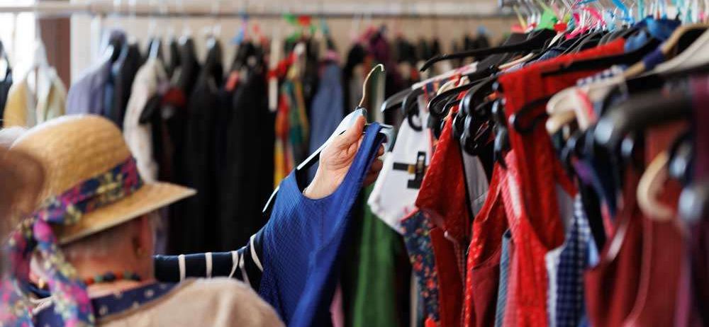 Eine Person steht vor einem Kleiderständer und betrachtet Kleidungsstücke an Kleiderbügeln, trägt einen Strohhut und eine Halskette.