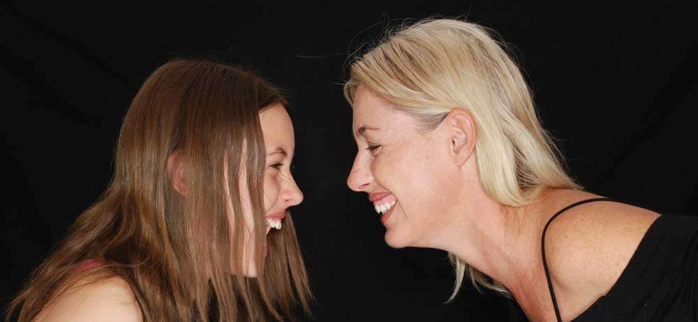 Zwei Frauen lachen und schauen sich an. Die Frau auf der linken Seite hat braune Haare und die Frau auf der rechten Seite hat blonde Haare. Sie tragen beide schwarze ärmellose Tops.