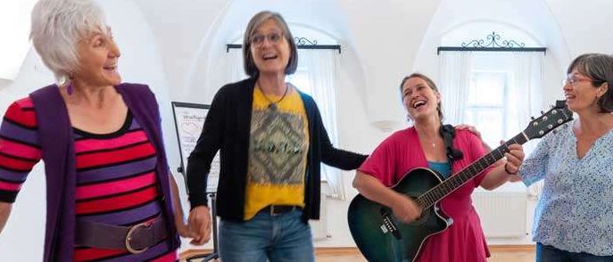 Vier Frauen, die lachen und tanzen, in einem Raum mit Holzboden. Eine spielt Gitarre. Ein Blumenstrauß steht im Vordergrund.